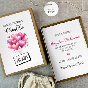 May include: A white card with pink and purple heart balloons and the text "HERZLICHEN GL&Uuml;CKWUNSCH Charlotte ABI 2024" is inside a brown box. A second white card with the text "DU HAST ES GESCHAFFTI! Herzlichen Gl&uuml;ckwunsch ZU DEINEM ERFOLGREICHEN ANSCHLUSS UND ALLES LIEBE UND GUTE F&Uuml;R DEINE ZUKUNFT WIR SIND GANZ STOLZ AUF DICH Mara Papa und Merity" is inside a gold box.
