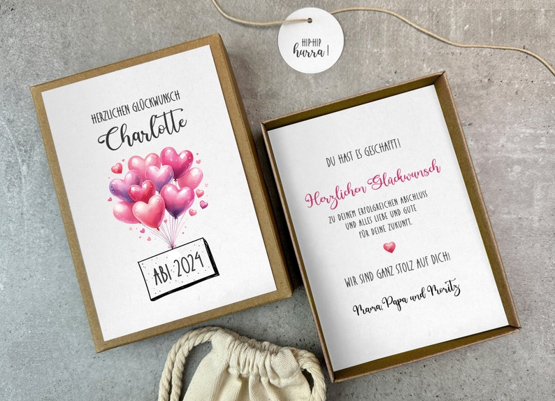 May include: A white card with pink and purple heart balloons and the text "HERZLICHEN GL&Uuml;CKWUNSCH Charlotte ABI 2024" is inside a brown box. A second white card with the text "DU HAST ES GESCHAFFTI! Herzlichen Gl&uuml;ckwunsch ZU DEINEM ERFOLGREICHEN ANSCHLUSS UND ALLES LIEBE UND GUTE F&Uuml;R DEINE ZUKUNFT WIR SIND GANZ STOLZ AUF DICH Mara Papa und Merity" is inside a gold box.