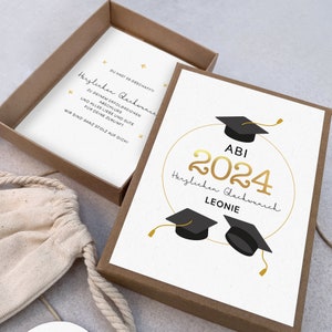 K&ouml;nnte beinhalten: Eine wei&szlig;e Karte mit goldener Schrift und schwarzen Abschlusskappen. Die Karte sagt "ABI 2024 Herzlichen Gl&uuml;ckwunsch Leonie".