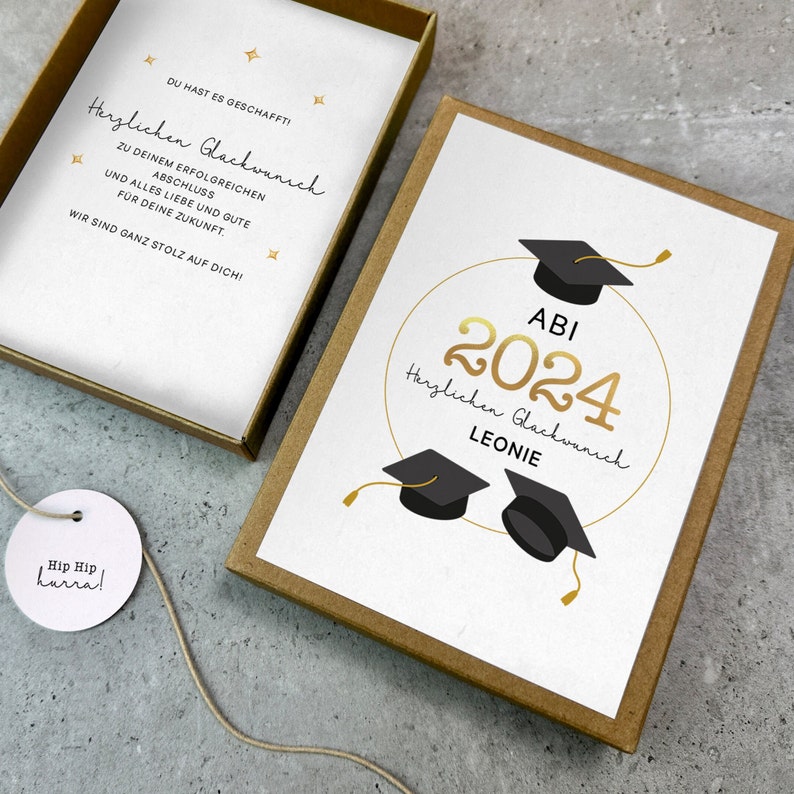 K&ouml;nnte beinhalten: Eine wei&szlig;e Karte mit goldener Schrift und schwarzen Abschlusskappen. Die Karte sagt "ABI 2024 Herzlichen Gl&uuml;ckwunsch Leonie".