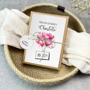May include: A brown card with a white background and a pink and purple balloon illustration. The card says "Herzlichen Gl&uuml;ckwunsch Charlotte" and "ABI 2024". A white circle tag with the text "HIP HIP hurra!" is attached to the card.