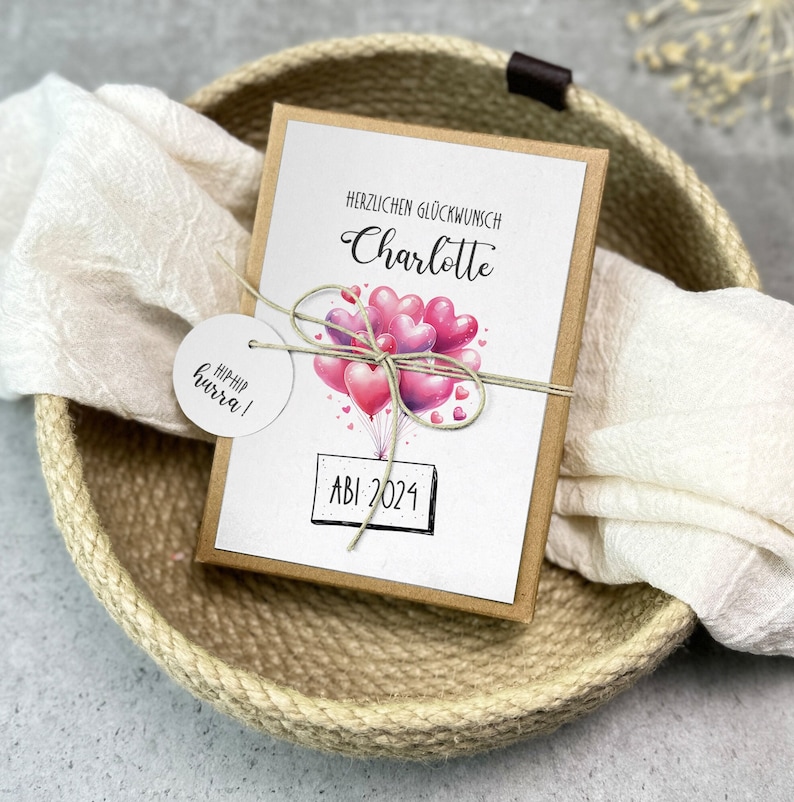 May include: A brown card with a white background and a pink and purple balloon illustration. The card says "Herzlichen Gl&uuml;ckwunsch Charlotte" and "ABI 2024". A white circle tag with the text "HIP HIP hurra!" is attached to the card.