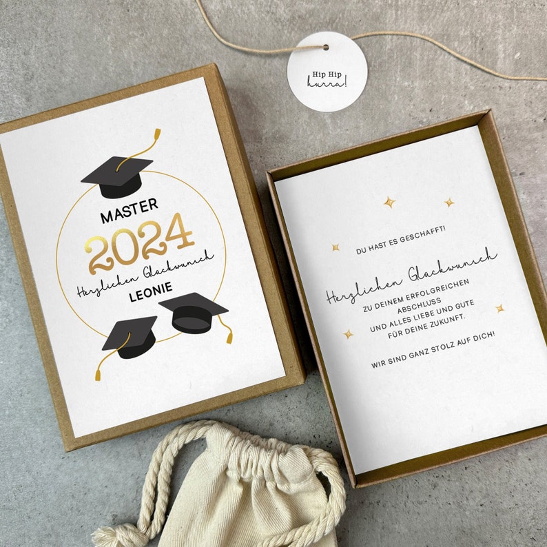 K&ouml;nnte beinhalten: Eine goldene und schwarze Abschlusskarte mit dem Text "MASTER 2024 Herzlichen Gl&uuml;ckwunsch LEONIE" und zwei schwarzen Abschlusskappen. Die Karte befindet sich in einer braunen Schachtel mit einem wei&szlig;en Etikett, auf dem "Hip Hip Hurra!" steht.