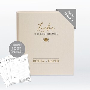 Könnte beinhalten: Ein beigefarbenes Leinenkochbuch mit dem Text "Liebe geht durch den Magen" und einer goldenen Gabel, einem Herzen und einem Messer. Das Kochbuch trägt außerdem den Text "Ronja & David" und ein Etikett mit der Aufschrift "Echtes Leinen".