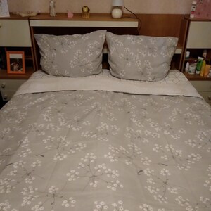 Peut inclure: Un lit gris avec une housse de couette à motif floral blanc et deux taies d'oreiller assorties. Le lit a une tête de lit en bois avec une petite lampe sur le dessus.