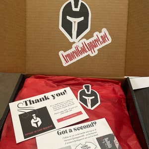 May include: A thank you card with a black and white illustration of a helmet and the text "Thank you!" and "Armor of God Apparel". A red tissue paper-filled box with a black and white helmet sticker and a second card with the text "Got a second?" and "Follow us on social media and tag us at @armorofgodapparellle on Instagram & armorofgodapparel.net on Facebook".