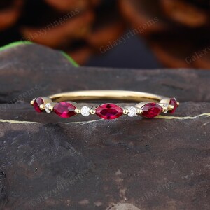 Peut inclure: Une bague en or avec des pierres précieuses rouges en forme de marquise alternant avec de petits diamants blancs ronds. La bague est posée sur un fond sombre et texturé. Le texte "galaxyringems" est visible en arrière-plan.
