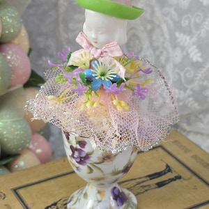 May include: A decorative porcelain egg cup with a floral design, topped with a doll head wearing a green hat and a pink bow. The doll is surrounded by a pink net skirt and colorful plastic flowers.