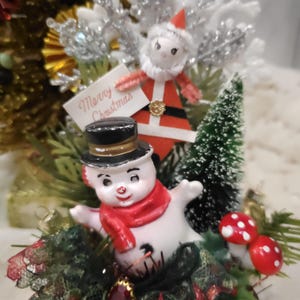 May include: A festive Christmas decoration featuring a snowman wearing a red scarf and a top hat, and a Santa Claus figure holding a "Merry Christmas" sign. The arrangement includes miniature trees and red and white mushroom ornaments.