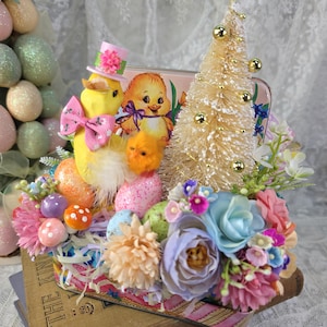 May include: An Easter-themed decorative display featuring a yellow chick wearing a pink hat and bow, a small gold tree, and colorful eggs and flowers. The display is set on a vintage book, creating a charming spring scene.