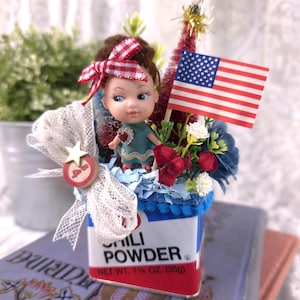 May include: A patriotic doll arrangement in a chili powder tin. The doll has a red and white bow, a heart-shaped pendant, and is surrounded by flowers, an American flag, and a small Christmas tree. The tin reads "CHILI POWDER" and "NET WT. 1 1/4 OZ. (35g)".