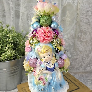 May include: A decorative ceramic figurine of a girl in a blue dress, holding a basket of flowers, topped with a floral arrangement and ornaments. The arrangement includes pink, blue, and green elements, creating a whimsical aesthetic. The figurine is placed on top of a stack of books.