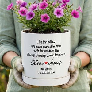 May include: A white ceramic planter containing purple petunia flowers. The planter features black printed text: Like the willow, we have learned to bend with the winds of life, always standing strong together. Olivia, a red heart, James, 12 years, 08.20.2014.