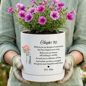 May include: A white ceramic flower pot containing purple petunia flowers. The pot features a printed illustration of a pink rose and text that reads: Chapter 70, Behind you are decades of memories that have shaped your story, EST. 1956.