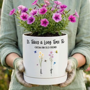 May include: A white ceramic planter containing vibrant purple petunia flowers. The pot features the black text "It Takes a Long Time To GROW AN OLD FRIEND" above two floral illustrations with the names "grace" and "emily" written in script.