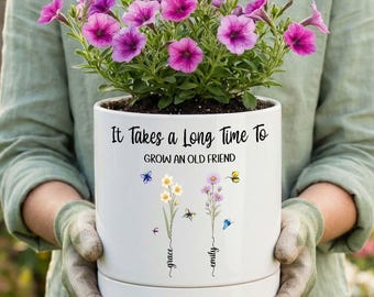 May include: A white ceramic planter containing vibrant purple petunia flowers. The pot features the black text "It Takes a Long Time To GROW AN OLD FRIEND" above two floral illustrations with the names "grace" and "emily" written in script.