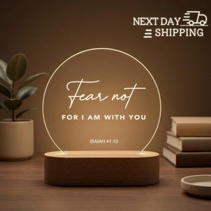 May include: A wooden-based, illuminated circular sign with the words "Fear not, for I am with you" in elegant script. The sign is lit with a warm glow, and the text "Isaiah 41:10" is at the bottom. The sign is on a wooden surface.