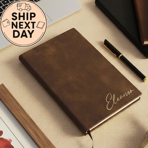 May include: A brown leather journal with the name "Eleanor" in cursive script. A black pen with gold accents rests beside the journal. The words "SHIP NEXT DAY" are in a circular graphic in the upper left corner. Other journals are in the background.