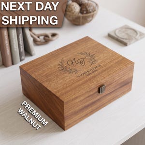 May include: A rectangular wooden keepsake box made of walnut with a hinged lid and a metal latch. The top of the box features an engraved design with the initials NT surrounded by a leaf wreath and the text NAOMI & THOMAS EST. 2018.