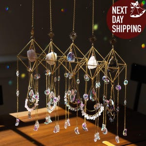 May include: Four hanging crystal suncatchers with gold geometric frames and chains. Each suncatcher features a different colored crystal and is adorned with iridescent beads and teardrop-shaped crystals. The suncatchers are suspended above a wooden table.