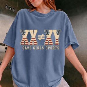 Könnte beinhalten: Ein blaues T-Shirt mit kurzen Ärmeln und einem Aufdruck mit dem Text XX ≠ XY sowie SAVE GIRLS SPORTS. Die Buchstaben sind mit einem abgenutzten US-Flaggenmuster in Rot, Weiß und Blau gefüllt und heben sich vom blauen Stoff ab.