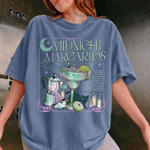 以下が含まれることがあります： 「MIDNIGHT MARGARITAS」の文字、ライムを添えたマルガリータグラス、コーヒーポット、紫色の魔女の帽子をかぶったカエル、黒猫のシルエット、キャンドル、小さな星が描かれたグラフィックプリント入りのスレートブルーのTシャツ。