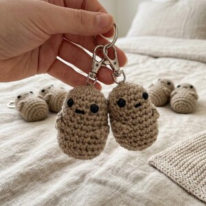 Może przedstawiać: Dwa szydełkowe breloczki w kształcie małych, beżowych ziemniaków z czarnymi guzikami i wyszywanymi ustami. Breloczki są przymocowane do srebrnych metalowych zapięć. W tle widocznych jest kilka innych szydełkowych przedmiotów w kształcie ziemniaków.