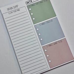 May include: A rectangular paper notepad with a white background featuring a date field, a Brain Dump section with lined space, and three colored boxes labeled 3 Things That I Have To Do in green, 3 Things I Could Do in blue, and 3 Things I Want To Do in pink.