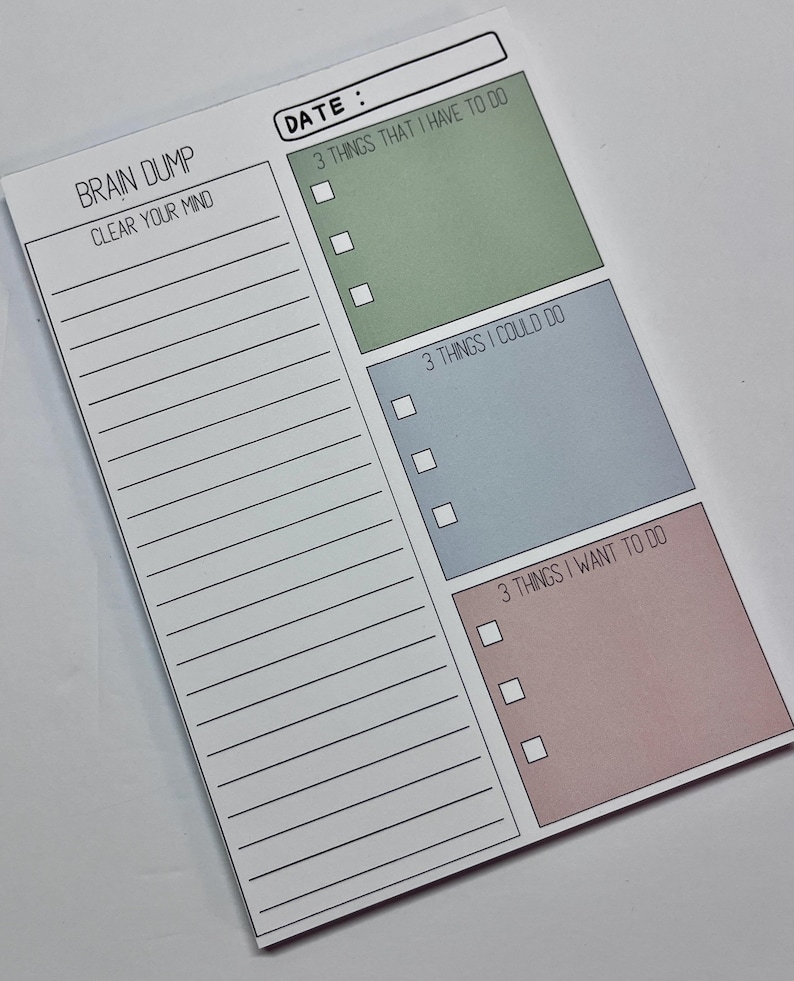 May include: A rectangular paper notepad with a white background featuring a date field, a Brain Dump section with lined space, and three colored boxes labeled 3 Things That I Have To Do in green, 3 Things I Could Do in blue, and 3 Things I Want To Do in pink.
