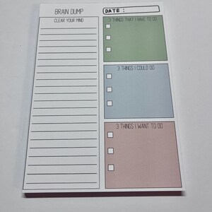May include: A rectangular notepad with a white background featuring the text BRAIN DUMP and DATE. The left side has lined space labeled CLEAR YOUR MIND. The right side has three colored boxes labeled 3 THINGS THAT I HAVE TO DO, 3 THINGS I COULD DO, and 3 THINGS I WANT TO DO, each with three checkboxes.