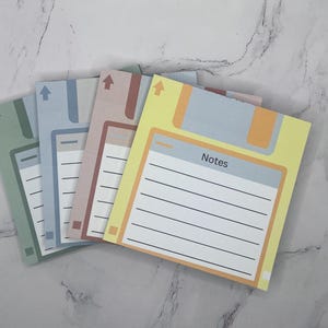 May include: A set of four square sticky note pads designed to resemble vintage floppy disks. Each pad features a different pastel color including yellow, pink, blue, and green, with a white lined writing area and the word Notes printed on top.