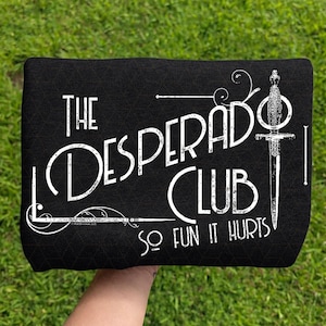 May include: A black fabric item featuring white printed text that reads The Desperado Club So Fun It Hurts. The design includes a vertical dagger illustration on the right side and decorative swirling line accents framing the text in a vintage style.
