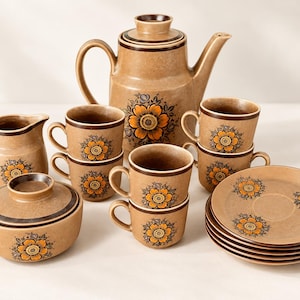 Op de afbeelding: Een koffieservies van keramiek met een beige gespikkelde glazuurlaag, een donkerbruine rand en een centraal oranje en bruin bloemmotief. De set bevat een koffiepot met deksel, een melkkan, een suikerpot met deksel, zes kopjes en zes schoteltjes.