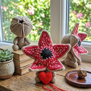 May include: Crocheted Demogorgon plush toys in various poses. The Demogorgons are primarily tan with red and white petal-like features. One has a red heart attached. A succulent plant and a wooden dice are also visible.