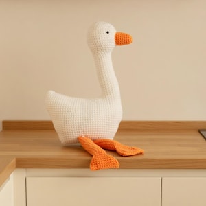 Könnte beinhalten: Eine handgefertigte weiße Häkelgans mit orangefarbenem Schnabel und Füßen. Die Gans sitzt auf einer Holzoberfläche. Die Häkelarbeit ist detailliert, und die Gans hat ein freundliches Aussehen. Ein charmantes Dekorationsstück.