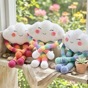 May include: Three handmade cloud-shaped plush toys with smiling faces and colorful legs. The clouds are white with pink cheeks and closed eyes. The legs are rainbow, orange, and blue. A small potted succulent sits nearby.