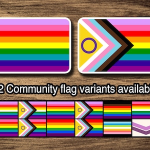 Può includere: Sono mostrate due versioni della bandiera Pride. La prima è la bandiera arcobaleno tradizionale con sei strisce: rosa, rosso, arancione, giallo, verde e viola. La seconda è una variante della bandiera Pride con un design a chevron e un cerchio viola con un contorno giallo. Il testo "12 varianti di bandiera della comunità disponibili" è sotto le bandiere.