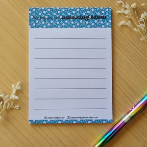 May include: A blue notepad with a white floral pattern and the text "Notes by an amazing Mum". The notepad has lined pages and a white border. The notepad is on a wooden surface with a rainbow pen.