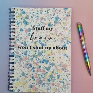 Op de afbeelding: Een spiraalgebonden notitieboek met een witte kaft met kleurrijke verfspetters en de tekst "Stuff my brain won't shut up about". Een regenboogkleurige pen ligt naast het notitieboek op een roze ondergrond. Schrijfwaren.