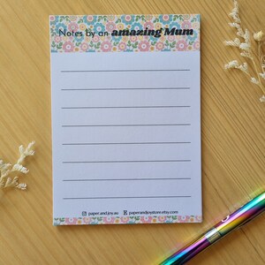 May include: A white notepad with a floral border and the text "Notes by an amazing Mum". The notepad has lined paper for writing notes. The notepad is on a wooden surface with a rainbow pen.