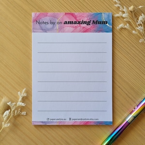 May include: A notepad with a pink and blue watercolour design and gold accents. The notepad has the text "Notes by an amazing Mum" at the top. The notepad is on a wooden surface with a rainbow pen.