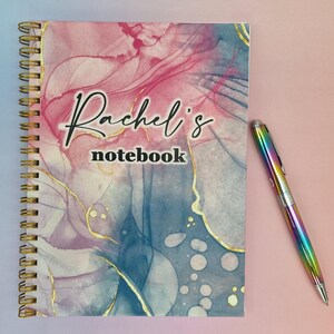 Op de afbeelding: Een roze en blauwe spiraalnotitieblok met gouden accenten en de tekst "Rachel's notebook" op de voorzijde.