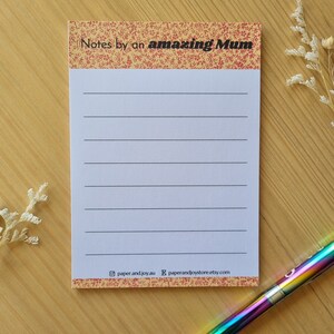 May include: A notepad with lined pages and the text "Notes by an amazing Mum". The top border features a red and yellow floral design. A rainbow pen lies on a wooden surface beside the notepad.