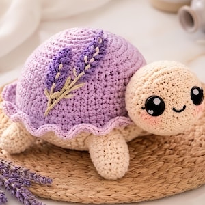 Puede incluir: Una tortuga de ganchillo hecha a mano en colores lavanda y crema. La tortuga tiene un caparazón lavanda con un diseño floral y una cabeza, patas y vientre de color crema. Está sobre una estera tejida.