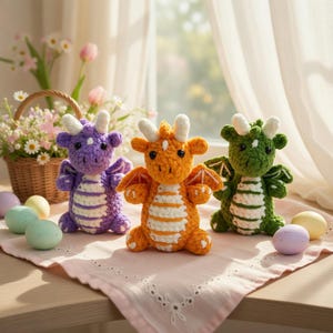 May include: Three handmade crochet dragons in purple, orange, and green, each with white accents and wings. They are standing on a pink cloth with Easter eggs and a basket of flowers in the background.