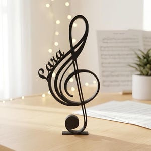 Könnte beinhalten: Eine schwarze, freistehende Skulptur in Form eines Violinschlüssels, in deren Design der Name Lara integriert ist. Das Objekt hat eine glatte, matte Oberfläche und steht aufrecht auf einem kleinen rechteckigen Sockel, was ein klares Musikmotiv zeigt.