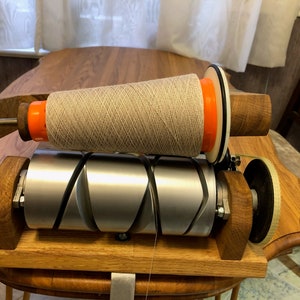 May include: A wooden yarn winder with a large spool of beige yarn. The winder has a silver metal roller and orange and white accents. The wooden base and supports are a light oak colour. The yarn is wound tightly on the spool.