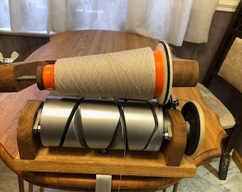 May include: A wooden yarn winder with a large spool of beige yarn. The winder has a silver metal roller and orange and white accents. The wooden base and supports are a light oak colour. The yarn is wound tightly on the spool.