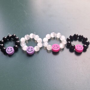 May include: Four beaded rings with smiley face accents. Two rings are black with purple smiley faces, and two are white with pink smiley faces. The rings are arranged on a green surface.