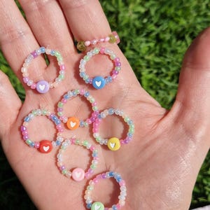 Könnte beinhalten: Ein Set aus sieben bunten Perlenringen, jeder mit einem weißen Herz-Anhänger. Die Ringe bestehen aus kleinen, schillernden Perlen in verschiedenen Farben, darunter Rosa, Blau, Grün, Gelb und Orange. Die Ringe werden an einer Hand mit einem bunten gewebten Armband gezeigt.